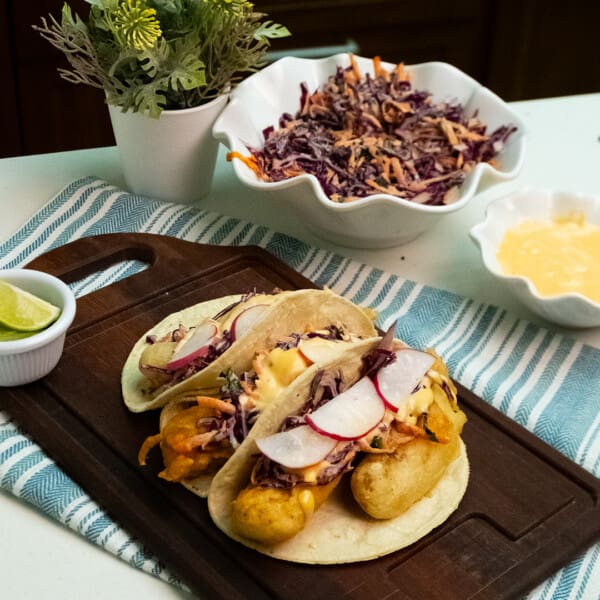 Three Baja-style fish tacos on a wooden cutting board, garnished with mint and lime wedges on the side.