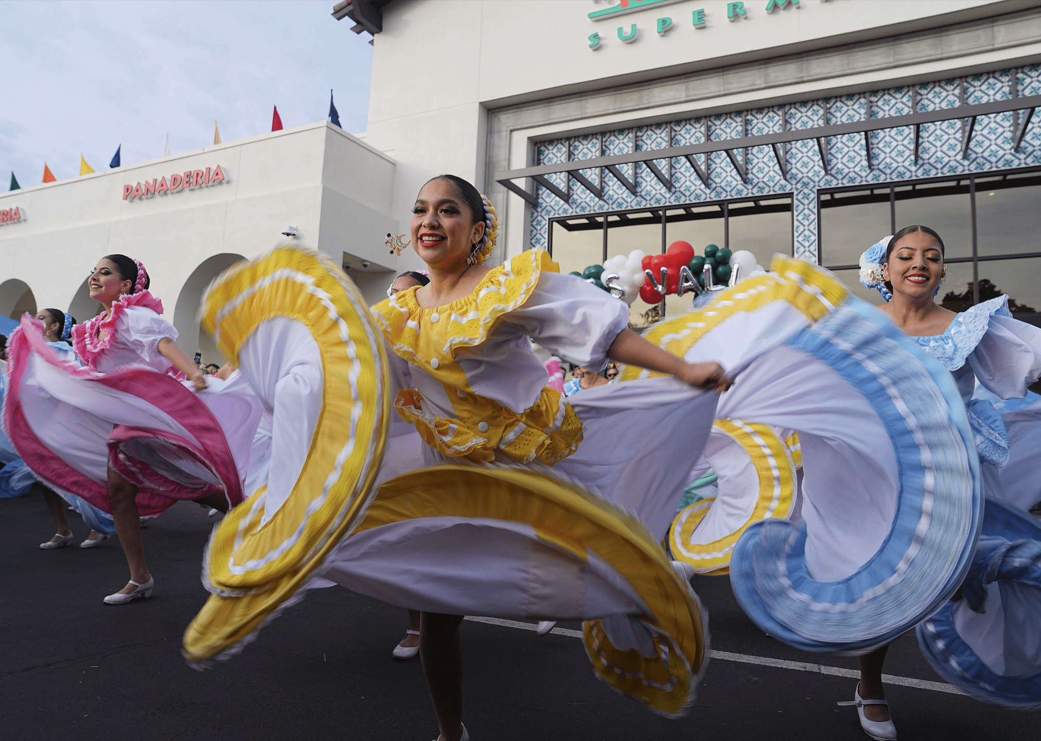 A group of folklorico dancers wearing vibrant yellow, baby blue, and pink traditional costumes performing on stage.
